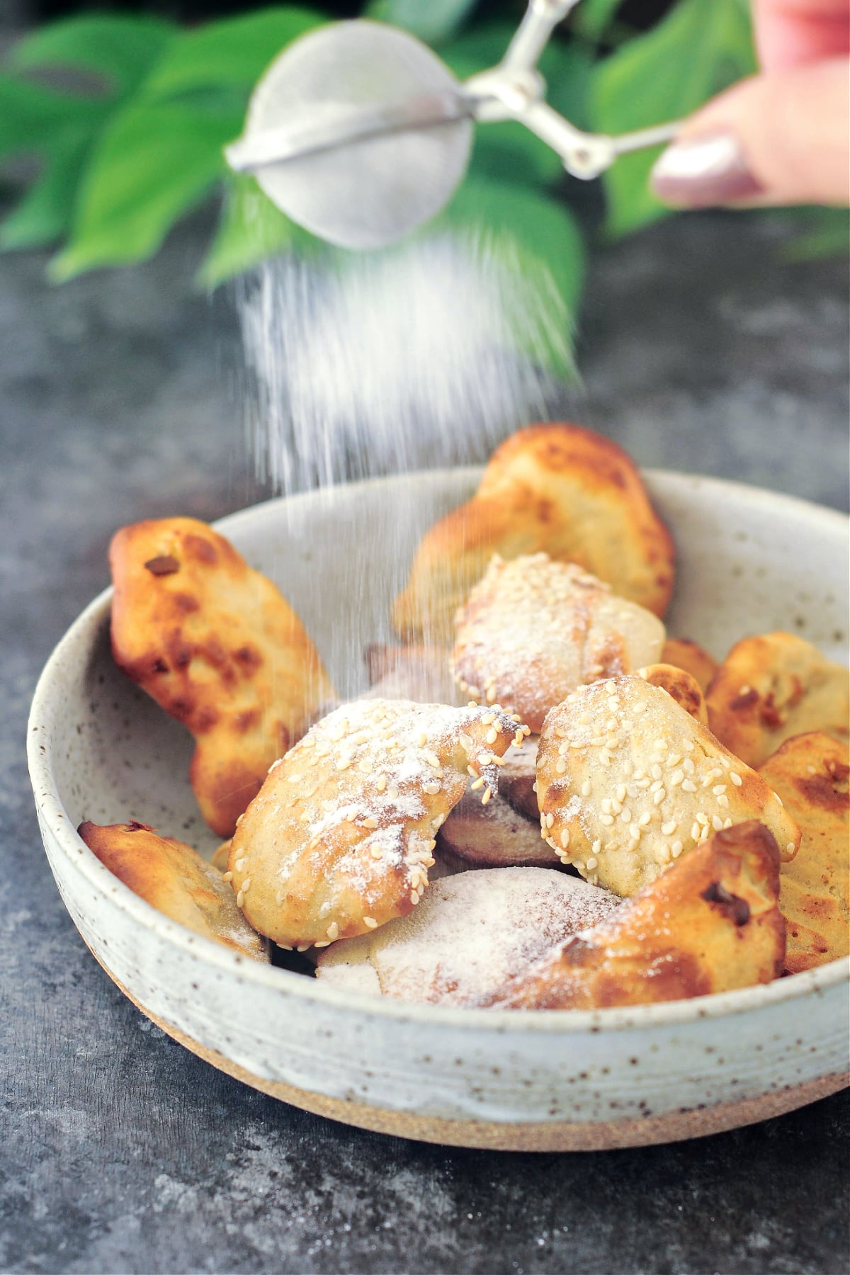 A hand holds a mesh strainer and dusts golden fried banana fritters in a shallow bowl.
