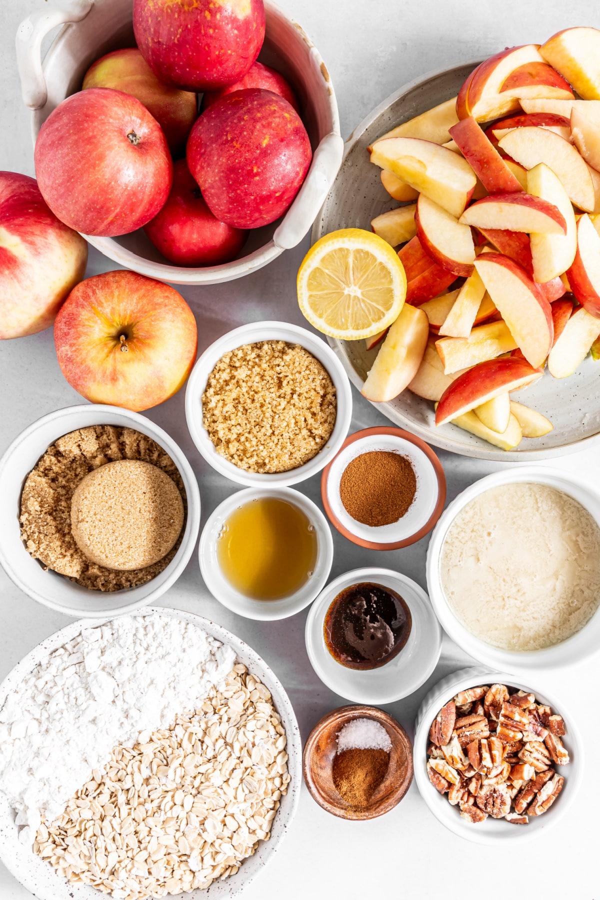 Overhead view of several small bowls of ingredients to make apple crisp: apples, sliced apples, lemon, sugars, spices, flour, oats, and pecan pieces.