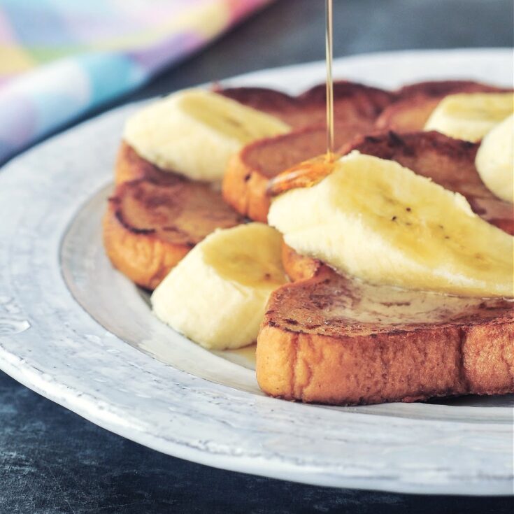 Close up of a stack of three slices of Baileys Irish cream French toast on a rustic white plate. French toast is garnished with sliced banana and maple syrup is being poured over the top.