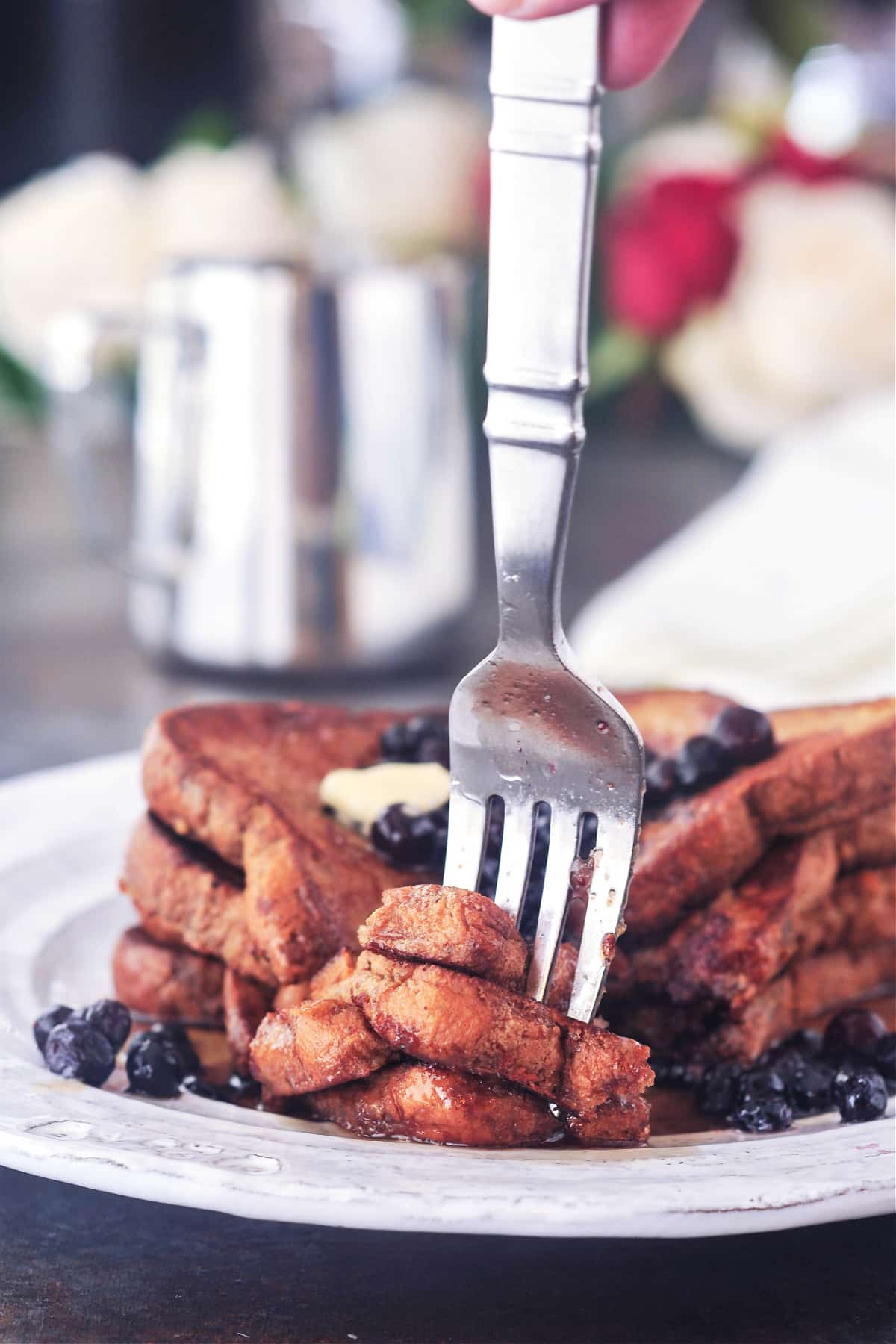 A stack of four slices of Baileys Irish cream French toast on a rustic white plate. French toast is garnished with blueberries and maple syrup, and a fork is picking up a bite cut from a corner.