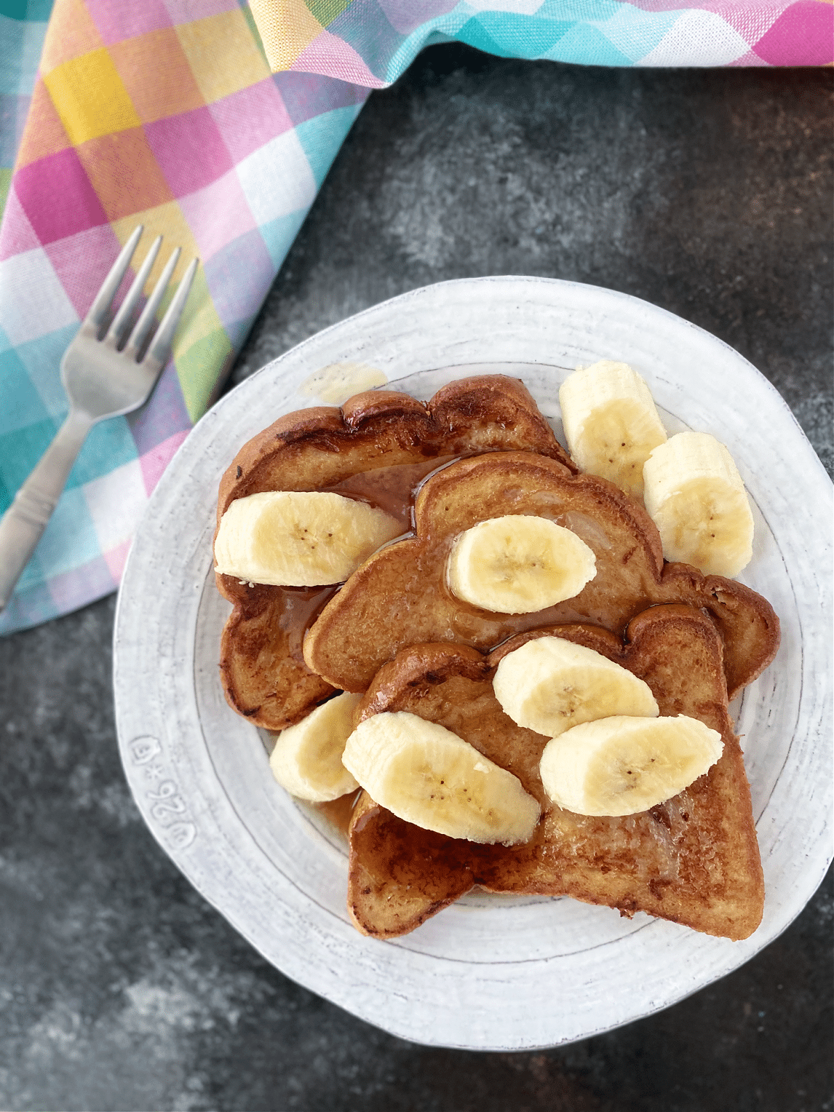 Overhead view of a stack of three slices of Baileys Irish cream French toast on a rustic white plate. French toast is garnished with sliced banana and maple syrup.