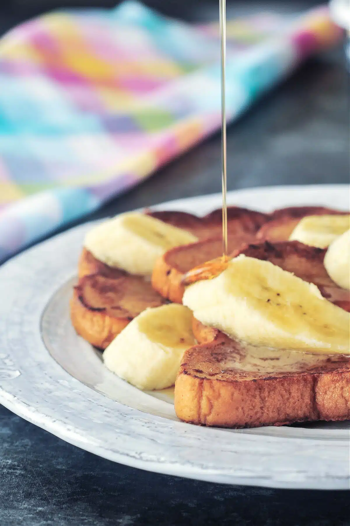 A stack of three slices of Baileys Irish cream French toast on a rustic white plate. French toast is garnished with sliced banana and maple syrup is being poured over the top.