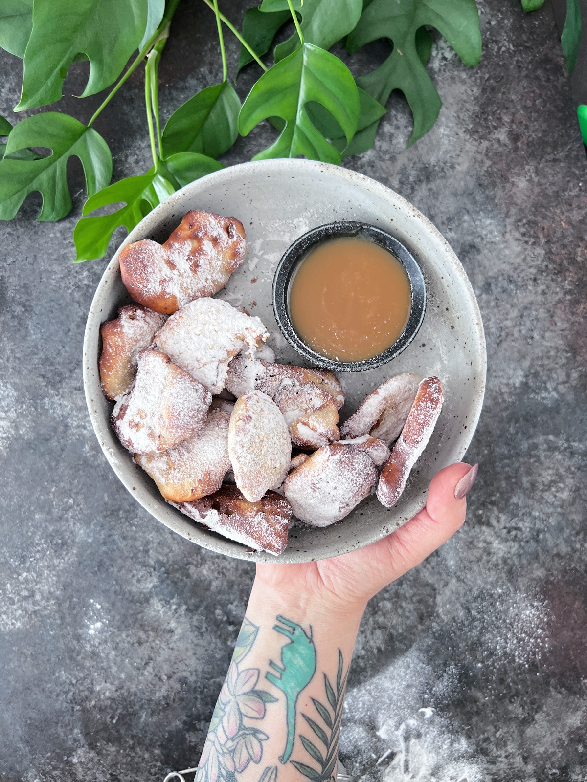 A hand holds a shallow bowl of banana fritters dusted with powdered sugar. A smaller bowl of caramel sauce sits in the fritter bowl. The arm has tattoos of foliage and a dinosaur.