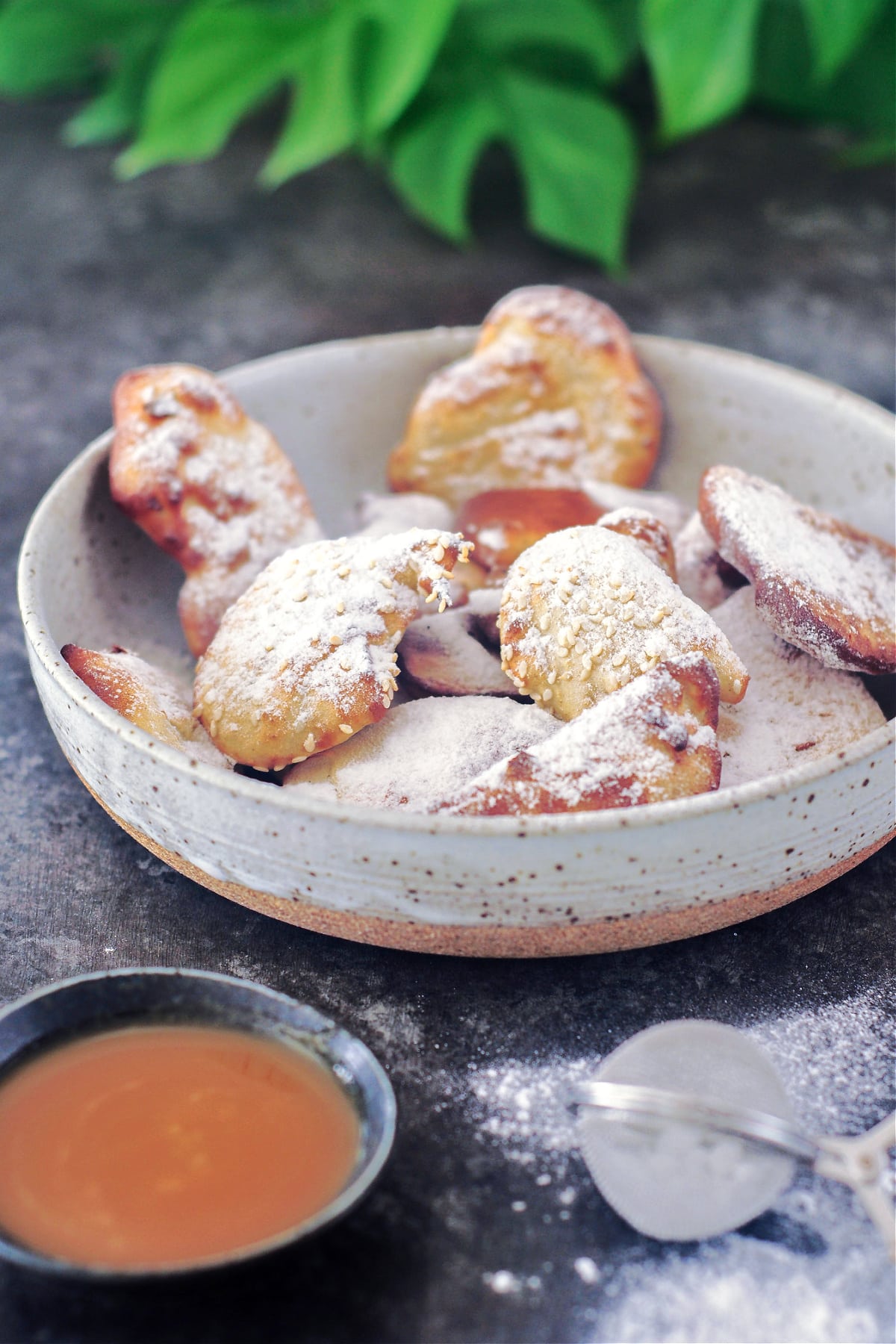 A shallow grey bowl of air fried banana fritters dusted with powdered sugar. A smaller bowl of caramel sauce and a mesh strainer for powdered sugar sit next to the bowl of fritters.