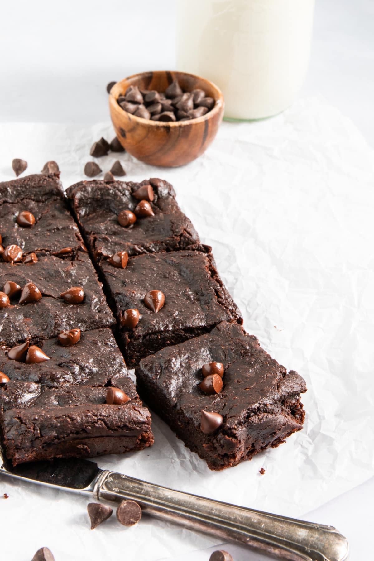 Six black bean brownies with chocolate chips cut into squares and sitting on a white surface next to a silver butter knife. A small bowl of chocolate chips in background, chocolate chips scattered around.