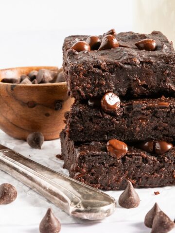 A stack of three black bean brownies with chocolate chips sitting on a white surface next to a silver butter knife. A small bowl of chocolate chips in background, chocolate chips scattered around.