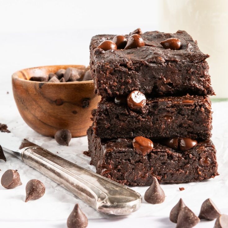 A stack of three black bean brownies with chocolate chips sitting on a white surface next to a silver butter knife. A small bowl of chocolate chips in background, chocolate chips scattered around.