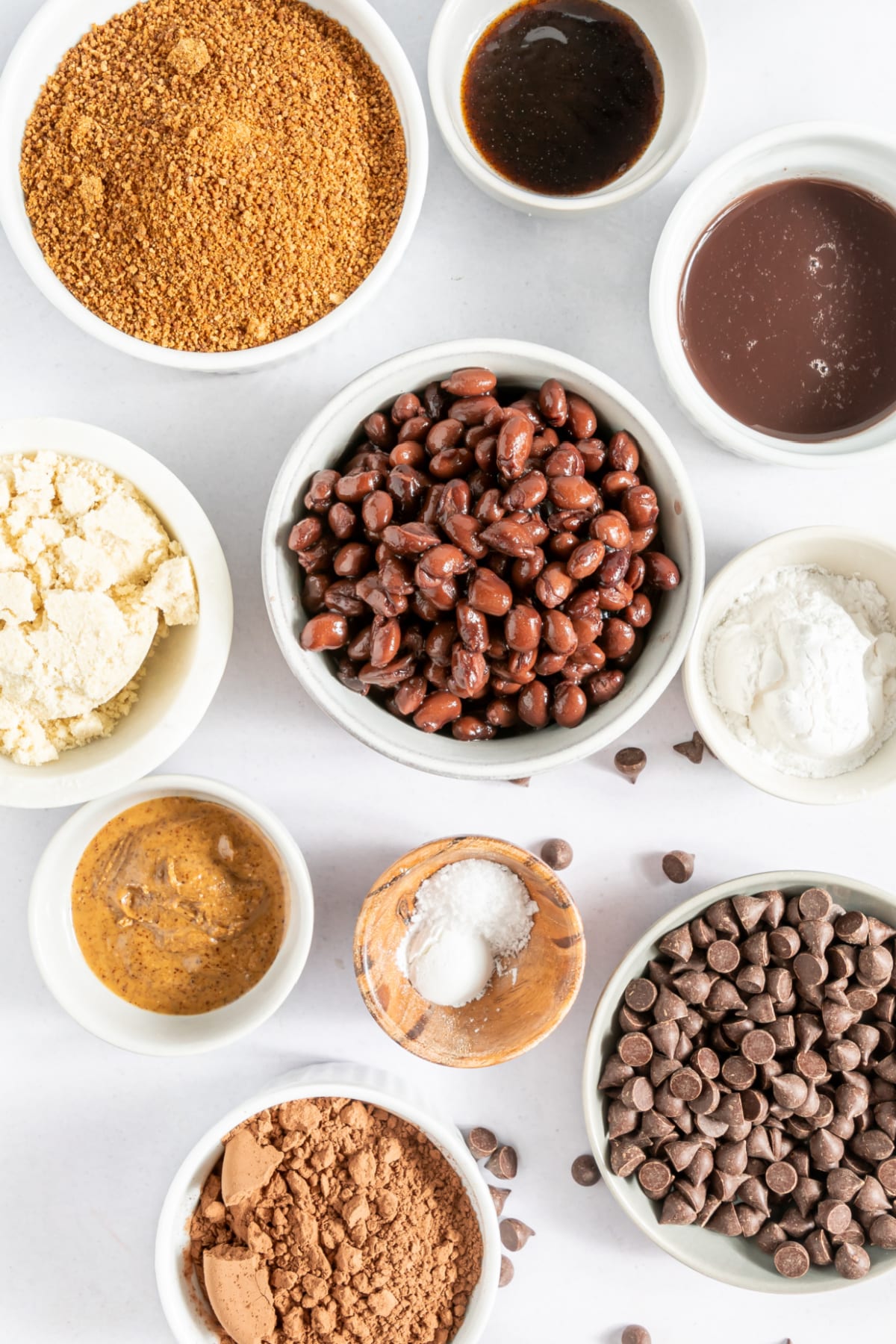 Overhead view of several small bowls of ingredients for black bean brownies: sugar, black beans, chocolate chips, cocoa powder. White bowls full of ingredients in various shades of brown on a white surface.
