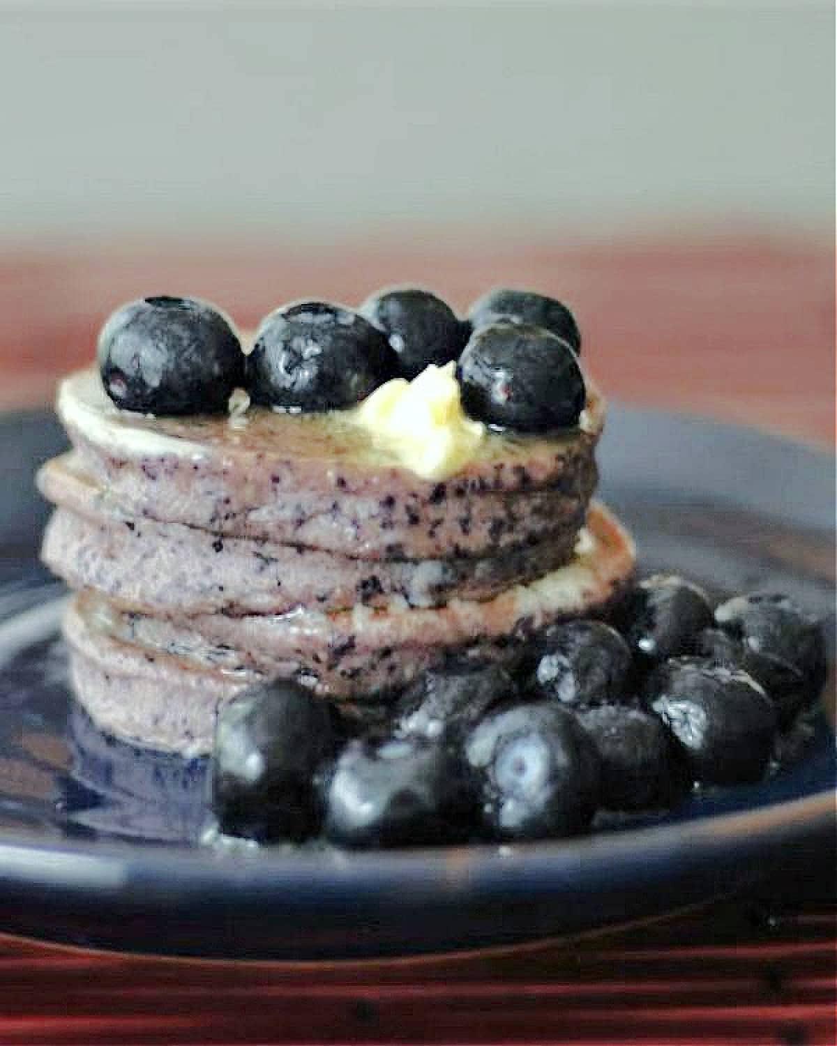 A stack of 4 pancakes that have fresh blueberries blended into the batter, so the pancakes are purple. Pancakes are on a dark blue plate and are garnished with melting butter, maple syrup, and plump juicy blueberries.