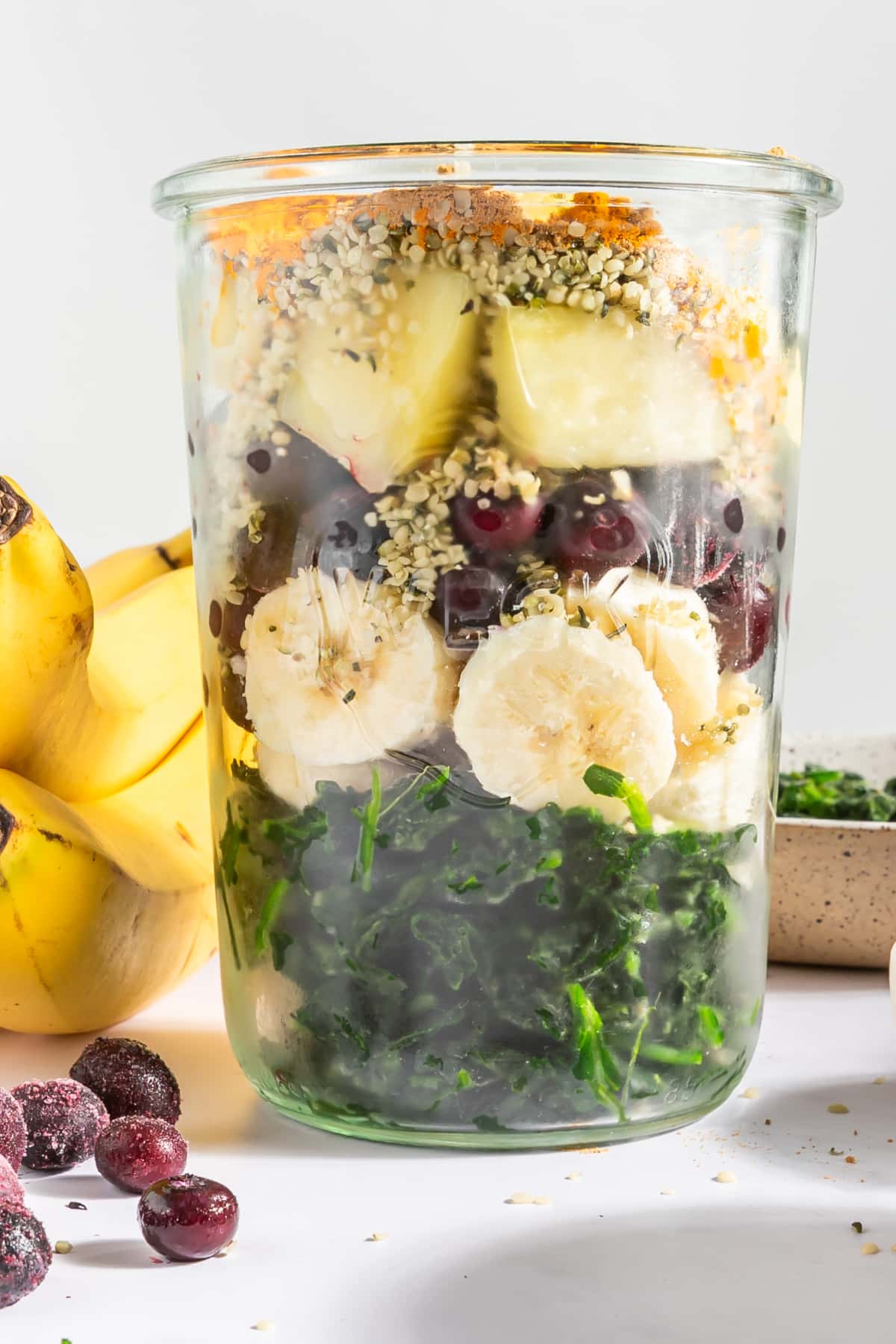 A large glass jar of smoothie ingredients for meal prepping: spinach, banana, pineapple, blueberries, hemp seeds, ground ginger, ground turmeric.