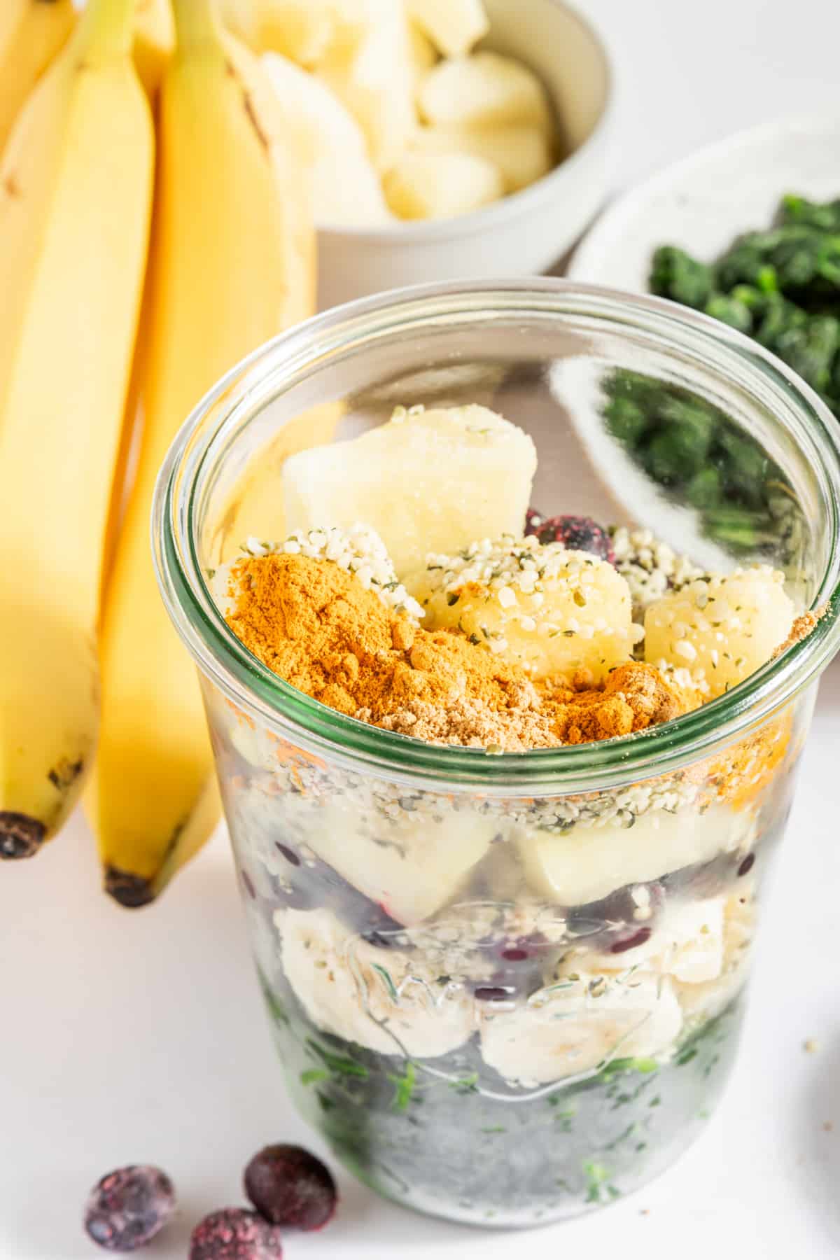 A large glass jar of smoothie ingredients for meal prepping: spinach, banana, pineapple, blueberries, hemp seeds, ground ginger, ground turmeric.