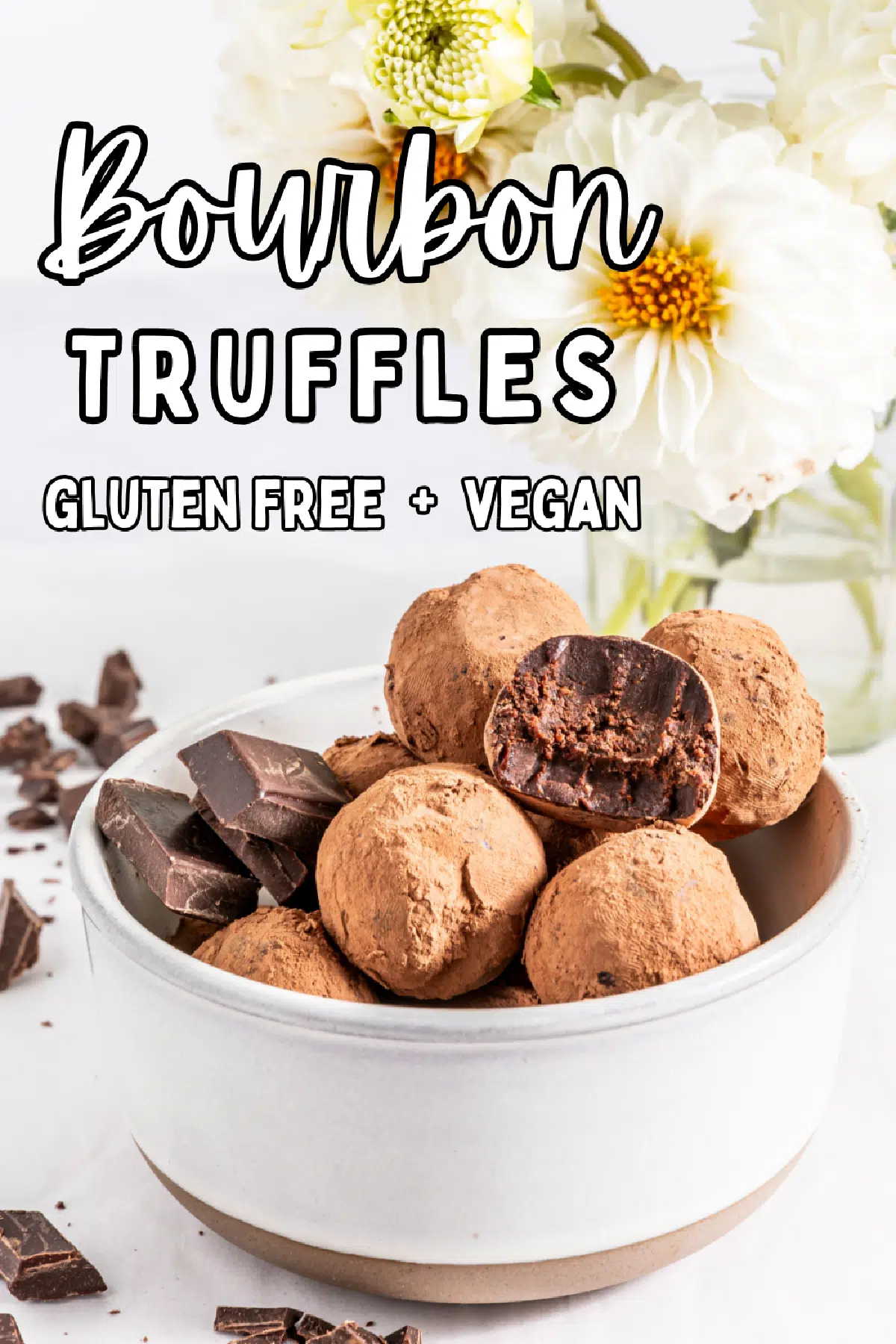 Chocolate truffles and a few squares of chocolate in a white bowl on a white tabletop, a white flower in a glass vase in background.