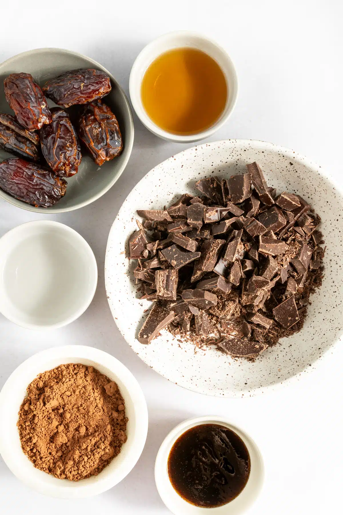 Several small bowls of ingredients to make bourbon balls: dates, bourbon, shopped chocolate, cocoa powder... Bowls are white and sit on a white surface.