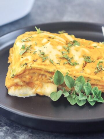 Close up side view of one serving of butternut sage lasagna on a matte black plate, with the white ceramic baking dish of lasagna blurred in the background. Lasagna is garnished with chopped fresh oregano and one whole sprig of oregano.