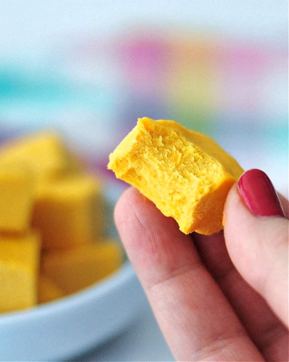 A hand holding a square of butternut toffee fudge, with the light blue shallow bowl of fudge blurred in background.