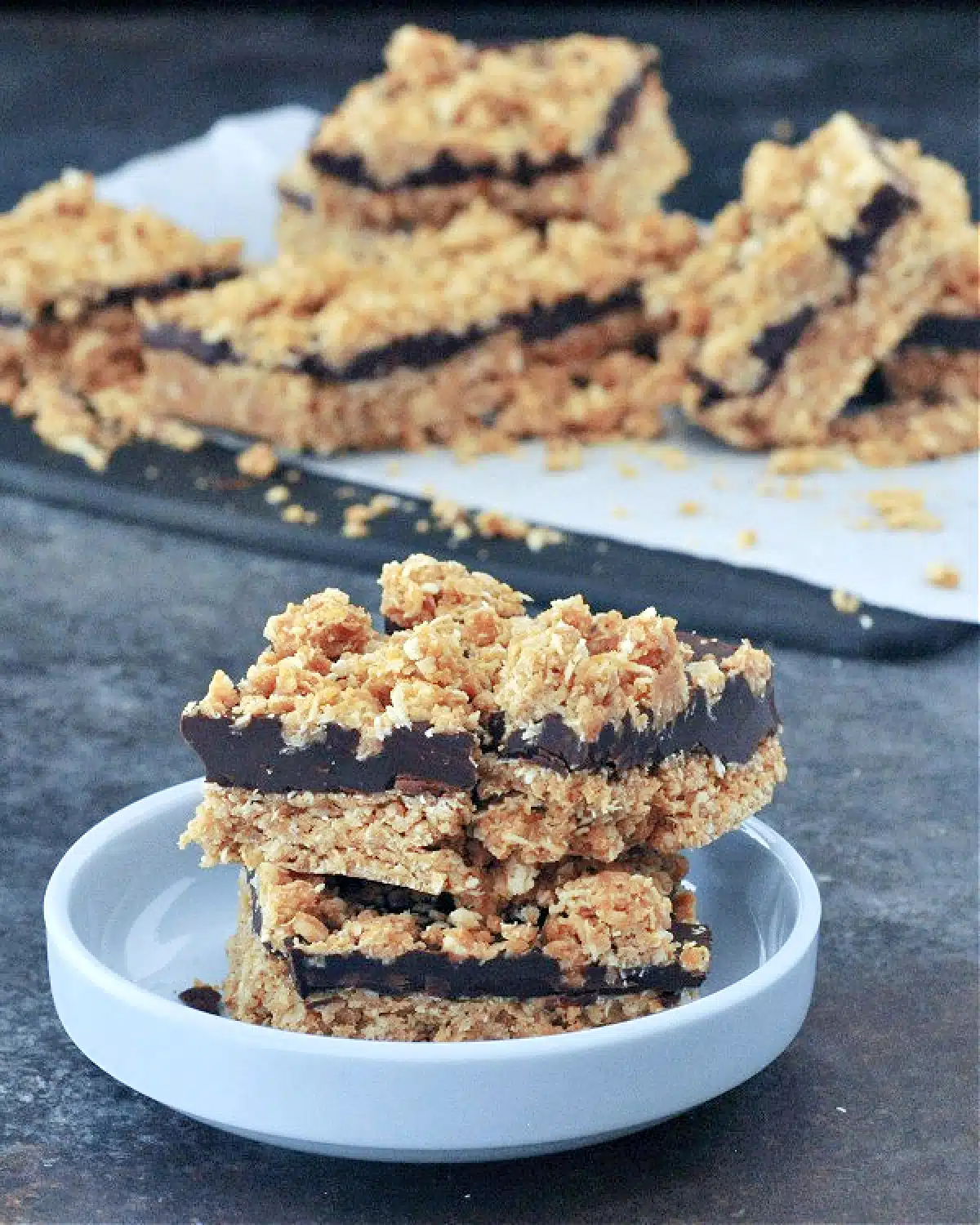 Two chocolate oat bars stacked in a shallow light blue dish. Several bars sit on parchment in background.