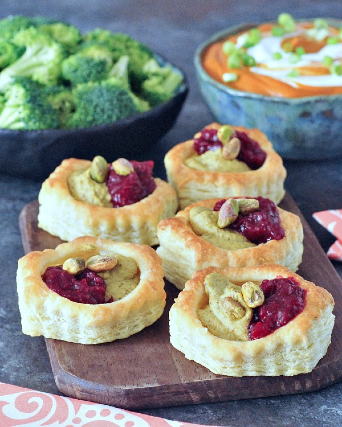 Bright pink and green garlic pistachio cranberry tarts on a wood serving board. A bowl of orange colored enchilada dip and a bowl of broccoli trees for dipping sit in the background.
