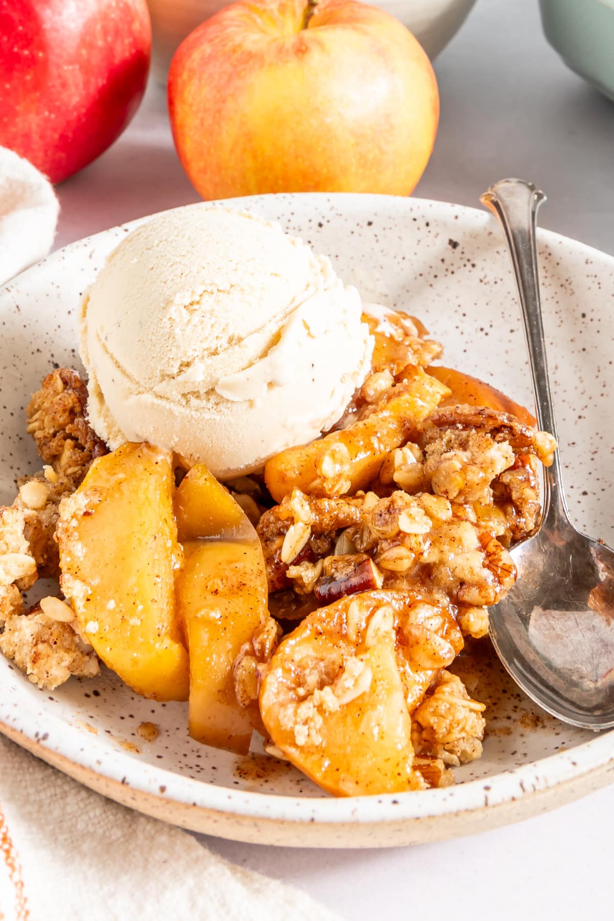 Close up of one serving of apple crisp and vanilla ice cream in a white bowl. The baking dish of apple crisp and a bowl of apples sits behind.