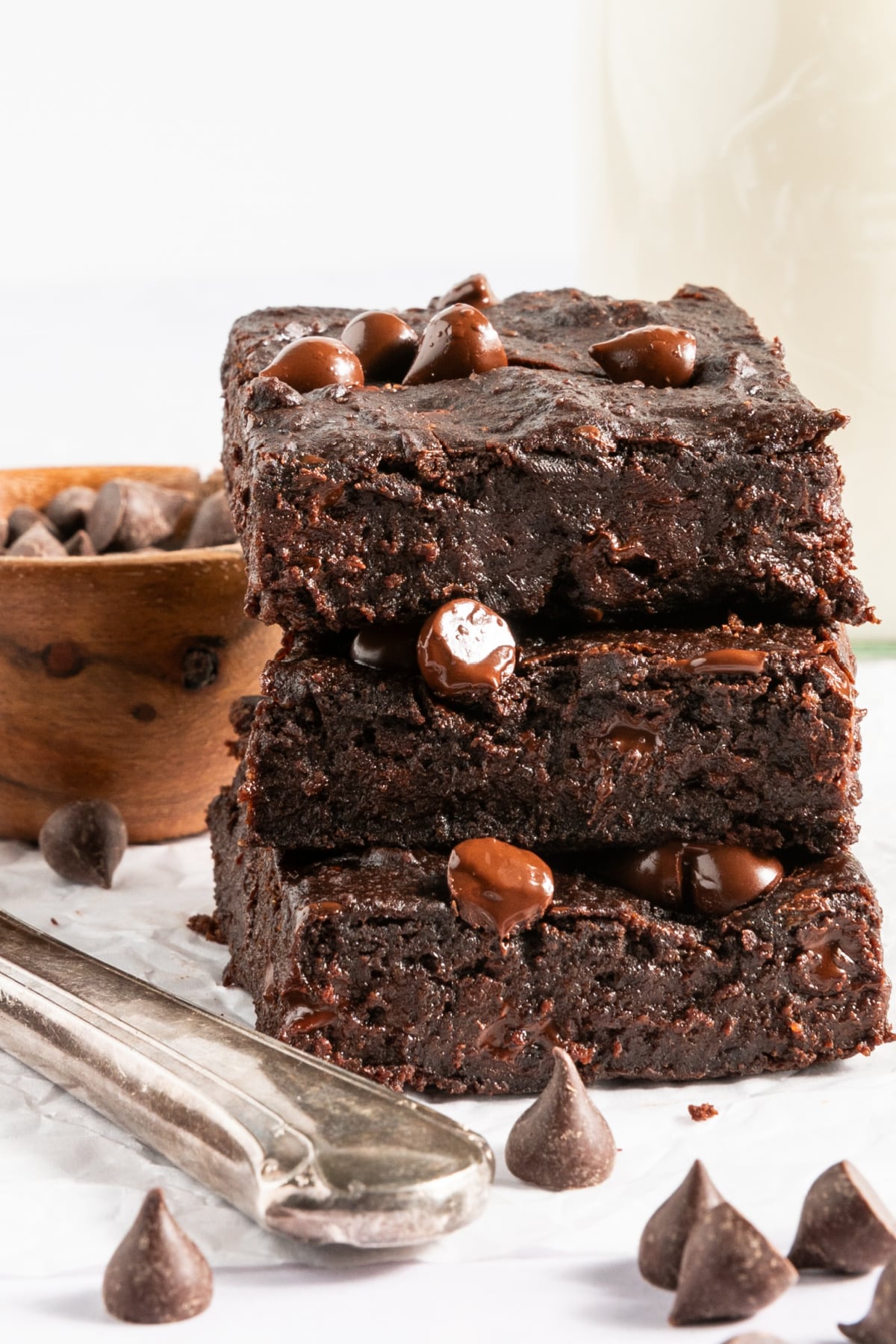 A stack of three gluten free black bean brownies with chocolate chips sitting on a white surface next to a silver butter knife. A small bowl of chocolate chips in background, chocolate chips scattered around.
