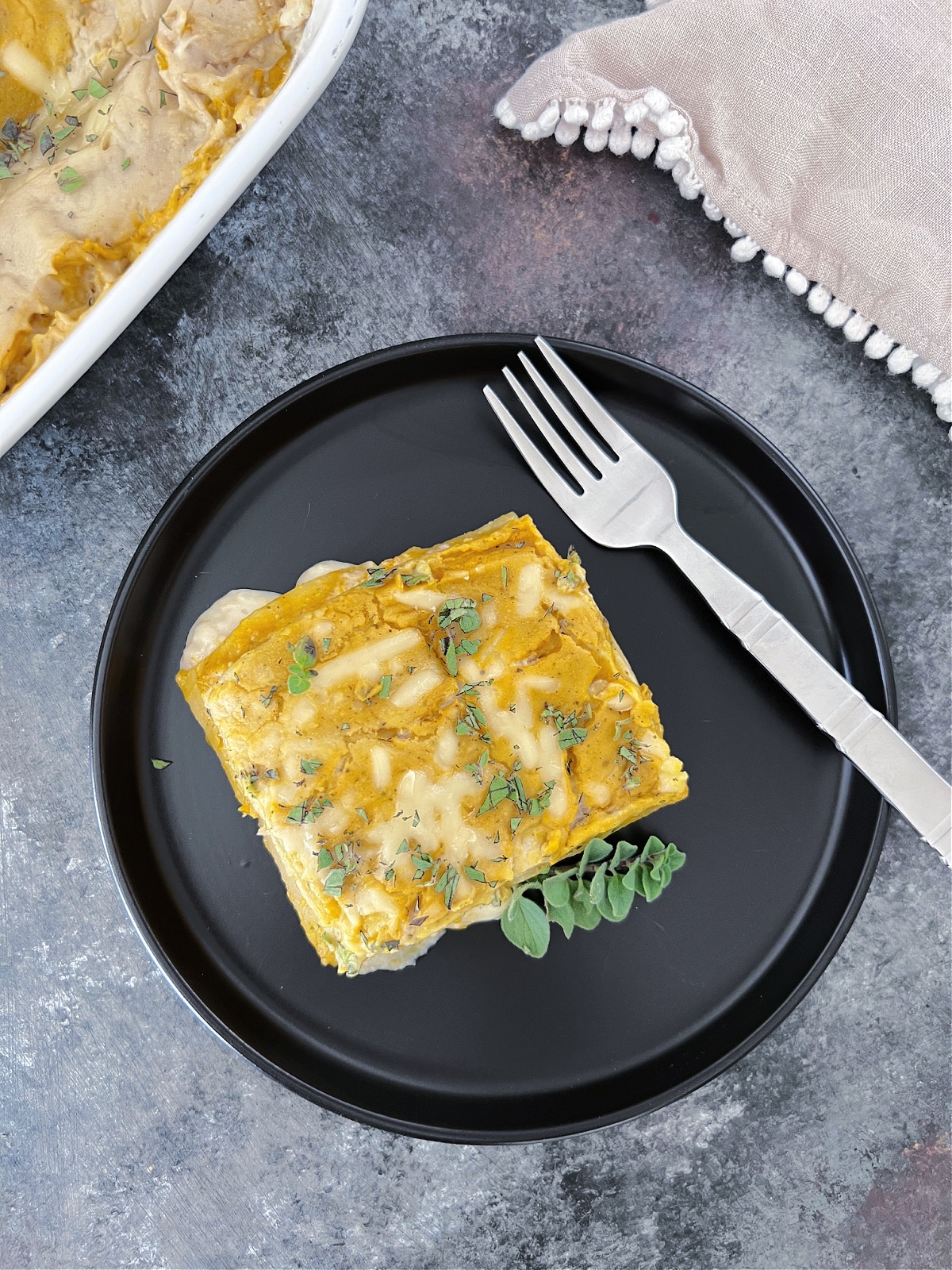 Overhead view of one serving of butternut sage lasagna on a matte black plate, with the white ceramic baking dish of lasagna blurred in the background. Lasagna is garnished with chopped fresh oregano and one whole sprig of oregano.