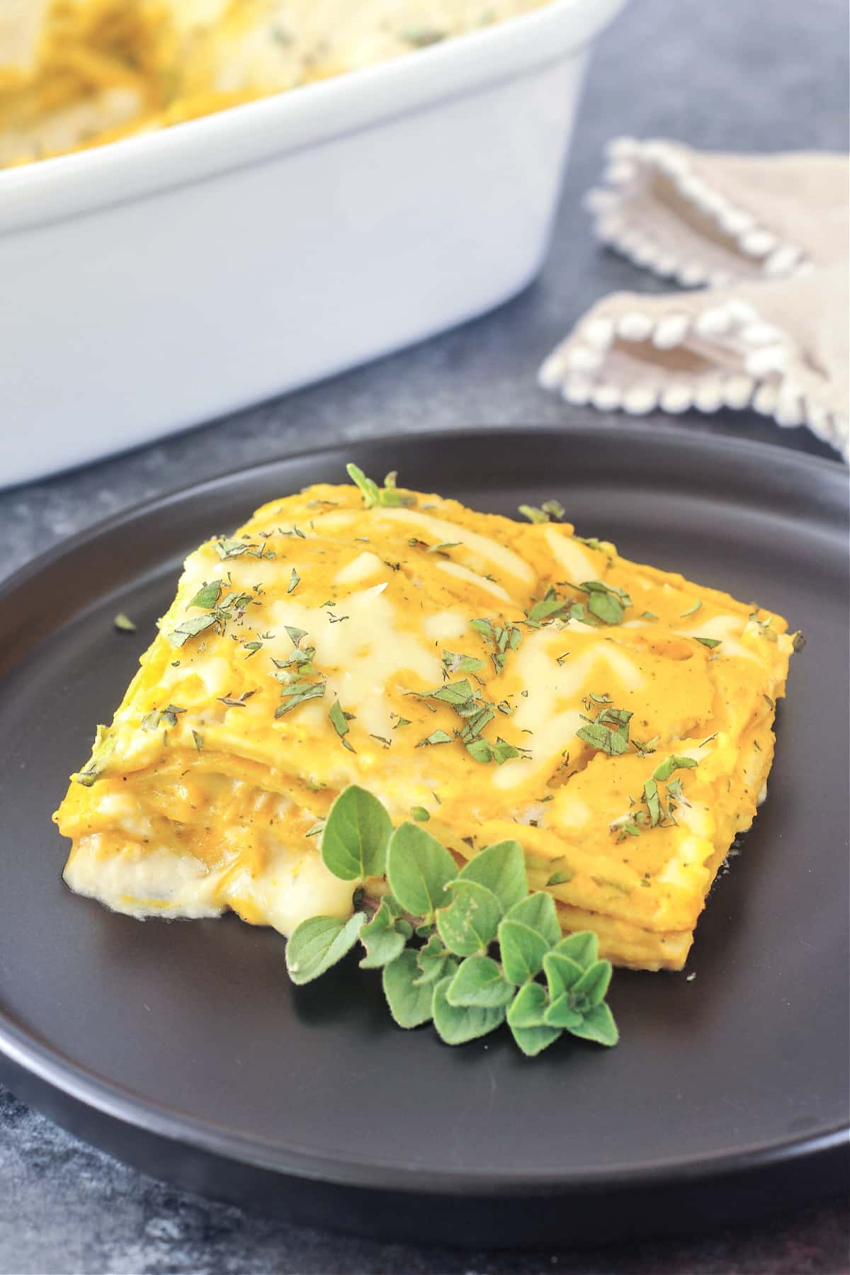 One serving of butternut sage lasagna on a matte black plate, with the white ceramic baking dish of lasagna blurred in the background. Lasagna is garnished with chopped fresh oregano and one whole sprig of oregano.