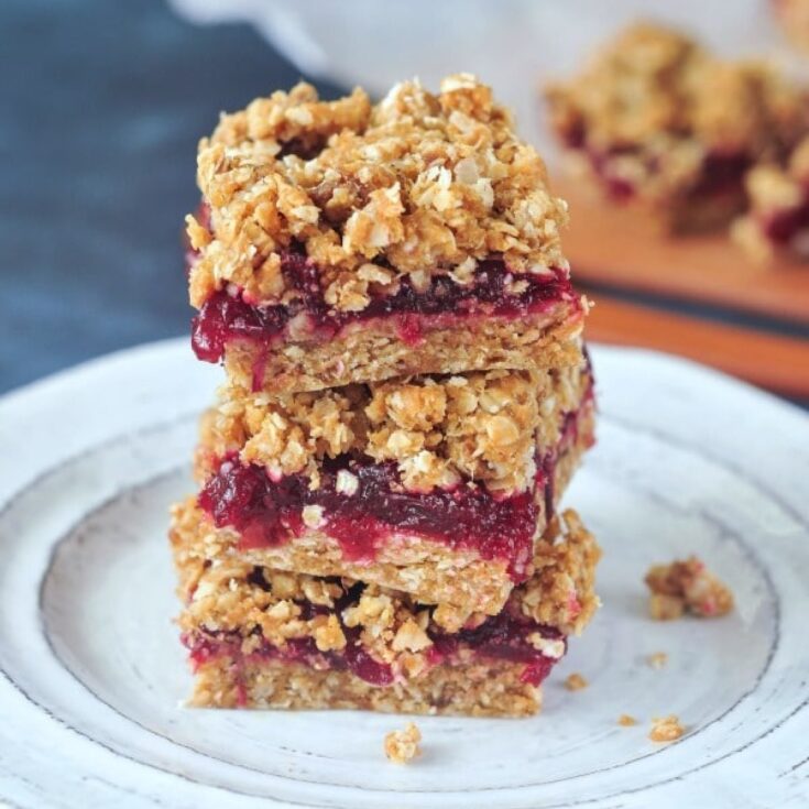 Close up of a stack of three cranberry bars on a rustic white plate against a dark blue marbled background.