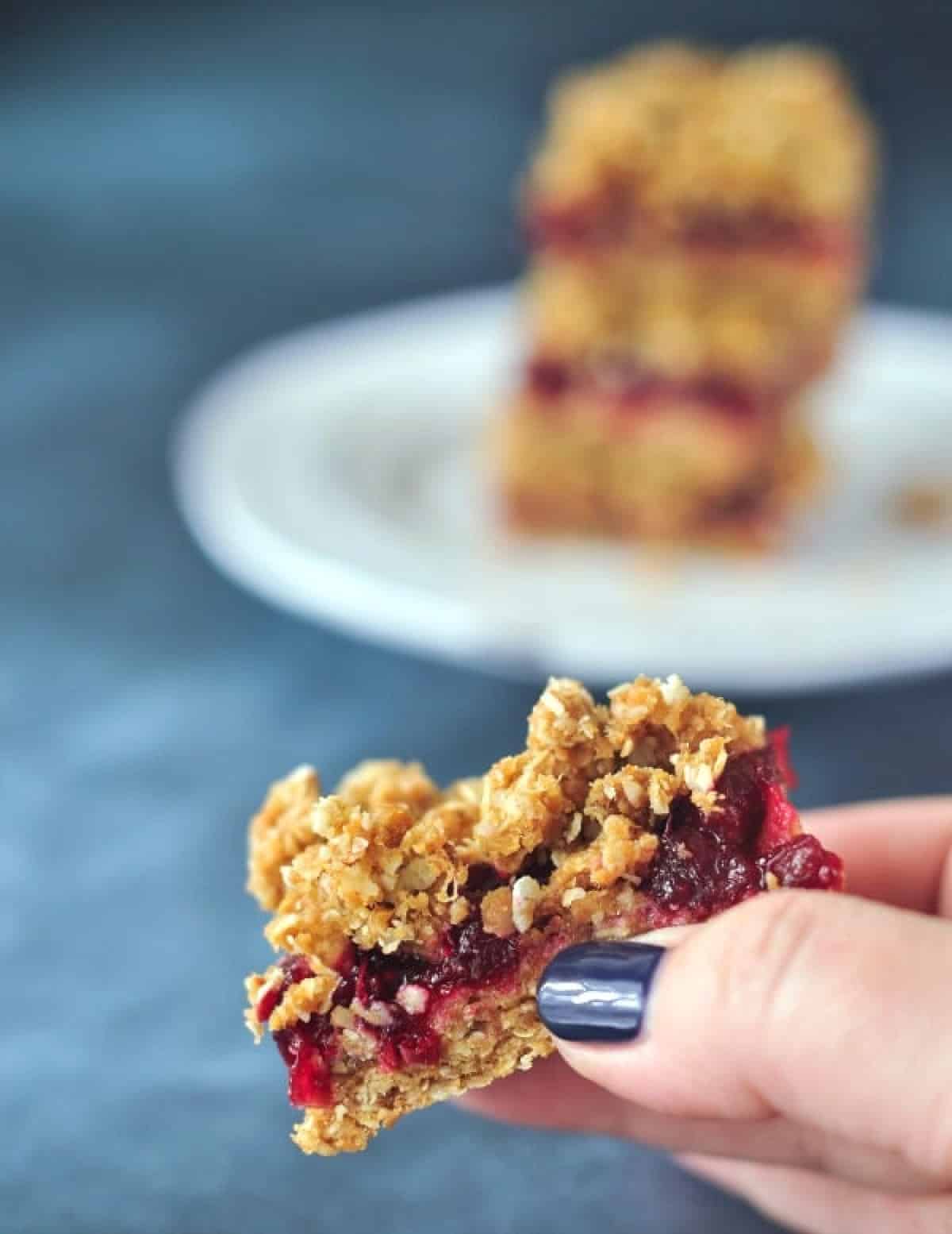 A hand holding one cranberry oat bar, with a stack on a plate blurred in background.