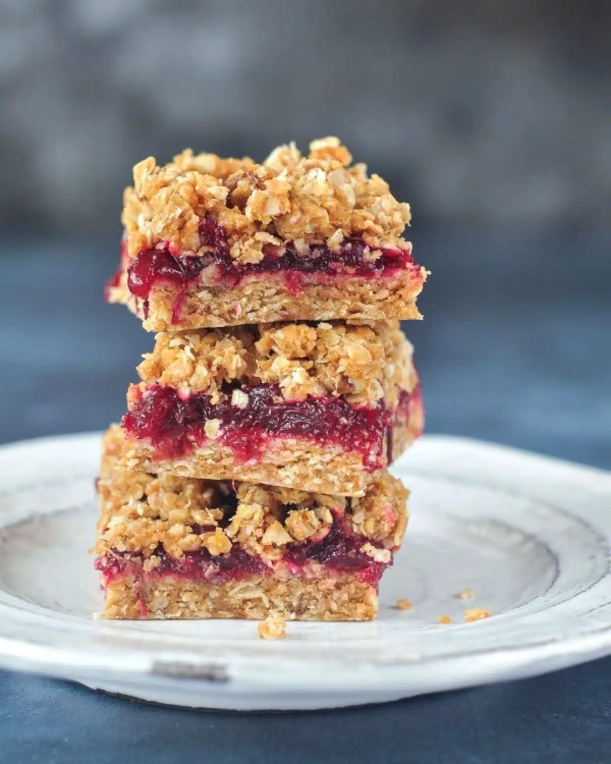 A stack of three cranberry oat no bake bars on a rustic white plate against a dark blue marbled background.