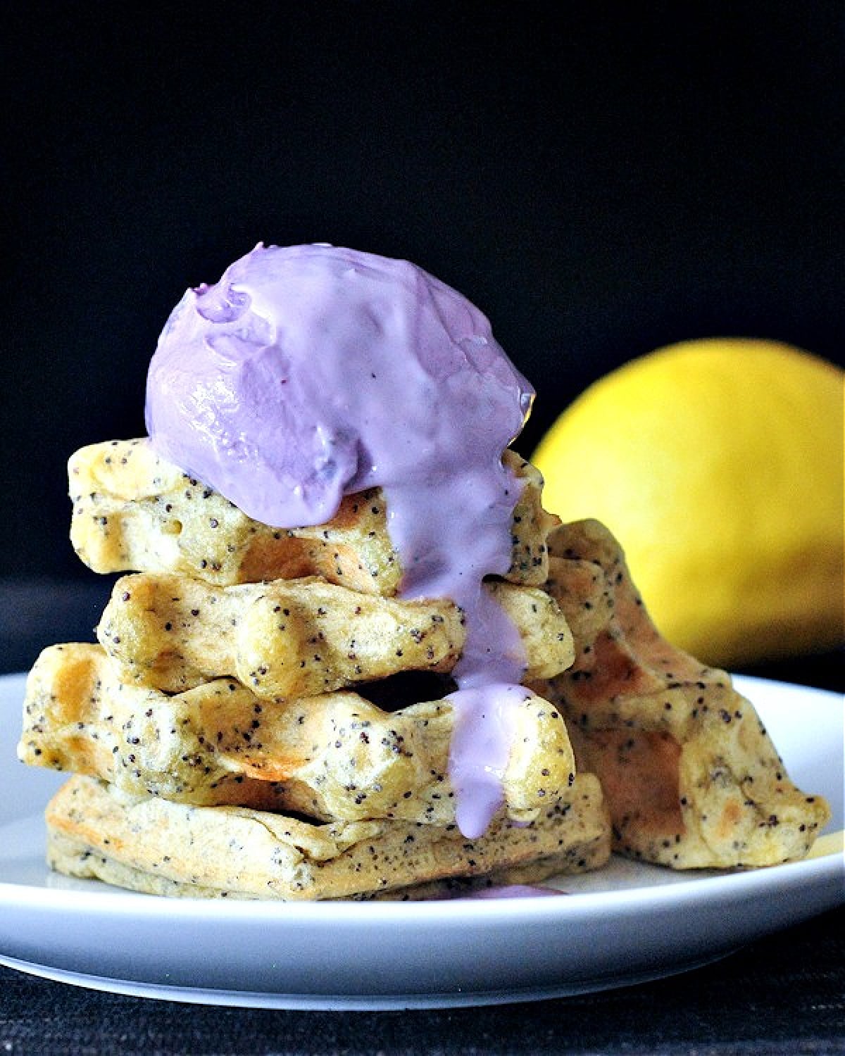 Side view of a stack of four golden yellow lemon poppyseed waffles on a white plate topped with a scoop of light purple blueberry yogurt. A whole lemon sits against a black background.