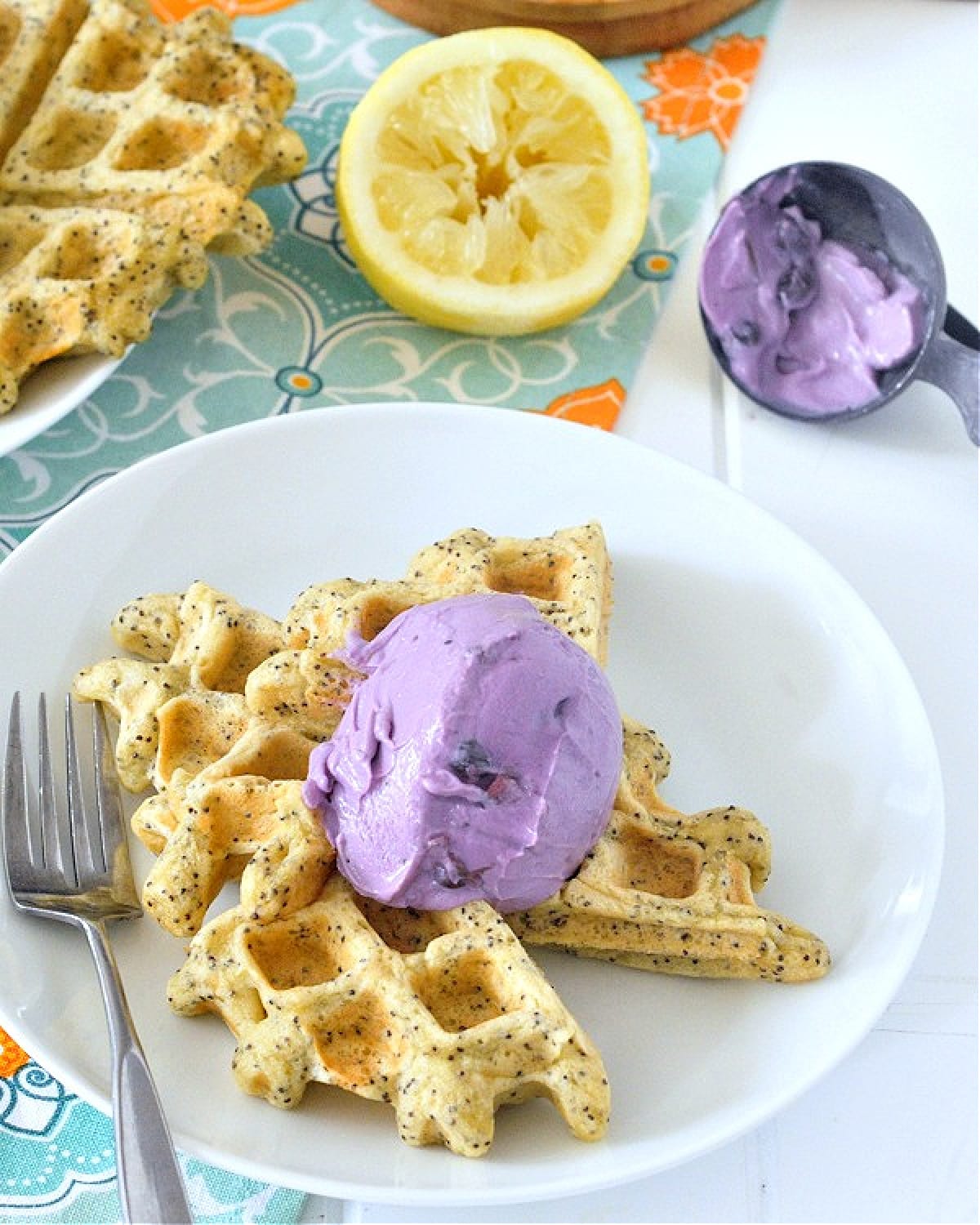 Golden yellow lemon poppyseed waffles on a white plate topped with a scoop of light purple blueberry yogurt. A fresh squeezed lemon half sits next to another white plate holding 2 more full Belgian style waffles, and an ice cream scoop with purple yogurt in it.