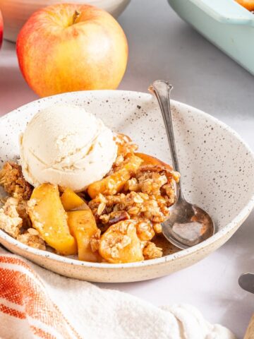 One serving of vegan apple crisp and vanilla ice cream in a white bowl. The baking dish of apple crisp and a bowl of apples sits behind.