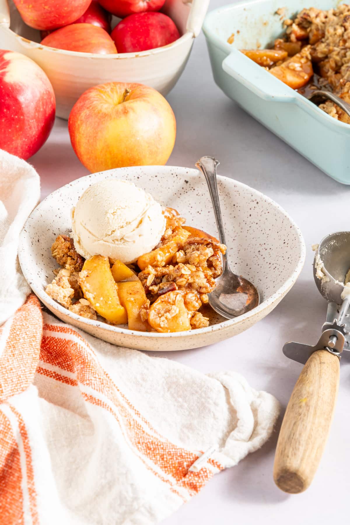 One serving of the gluten free apple crisp and vanilla ice cream in a white bowl. The baking dish of apple crisp and a bowl of apples sits behind.