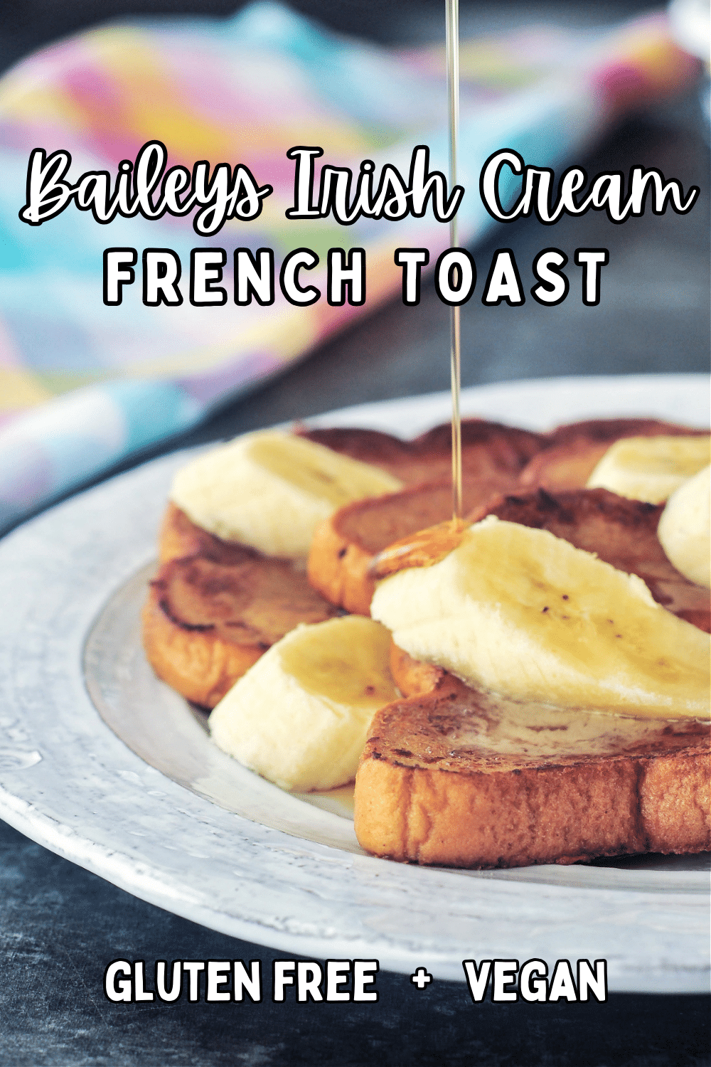A stack of three slices of Baileys Irish cream French toast on a rustic white plate. French toast is garnished with sliced banana and maple syrup is being poured over the top.