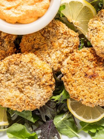 Golden brown vegan crab cakes and lemon slices on a bed of greens on a white platter, with a small white bowl of Thousand Island dressing.