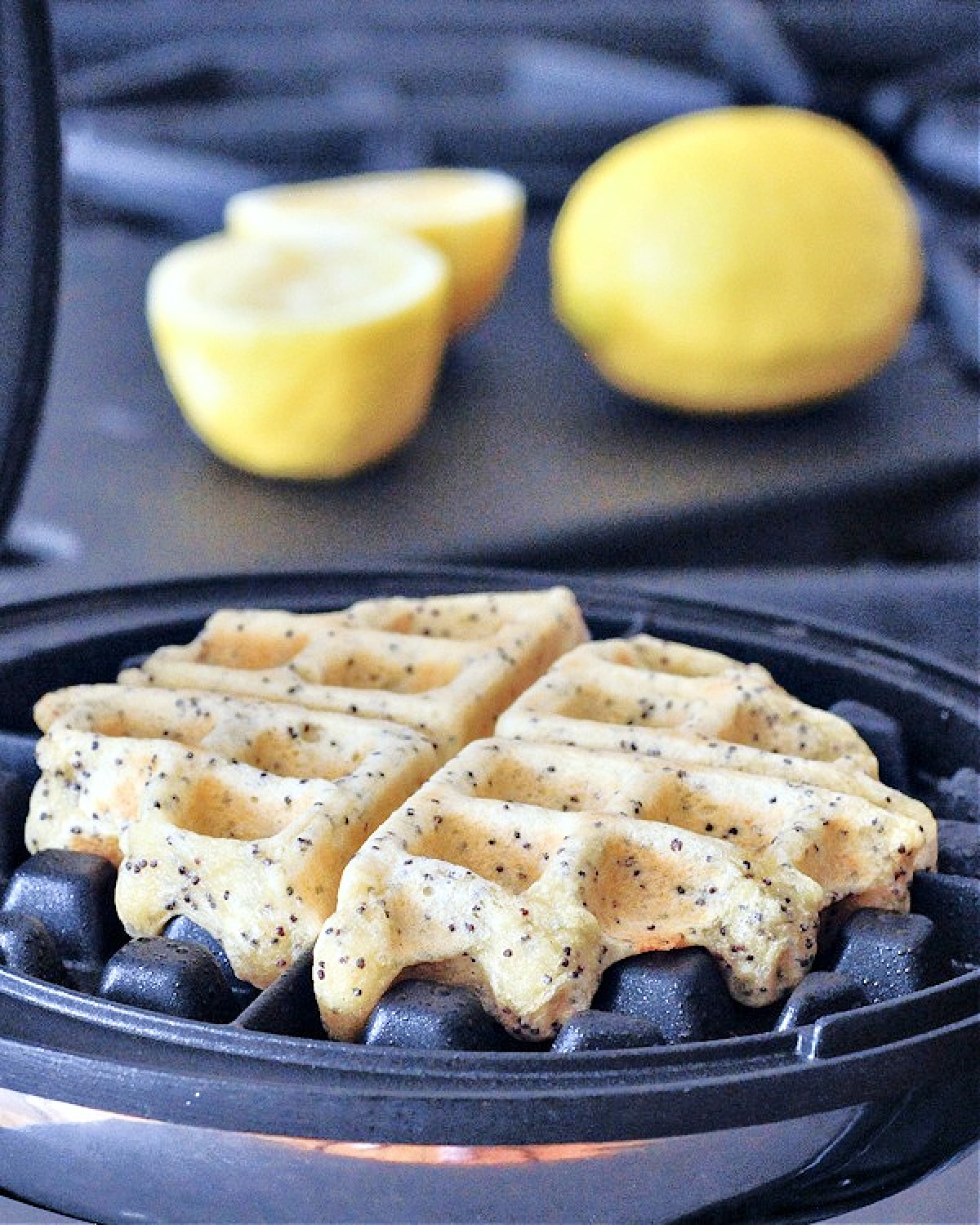 One full Belgian style lemon poppyseed waffle sits in the waffle maker. A whole lemon and one lemon cut in half sits blurred in the background.