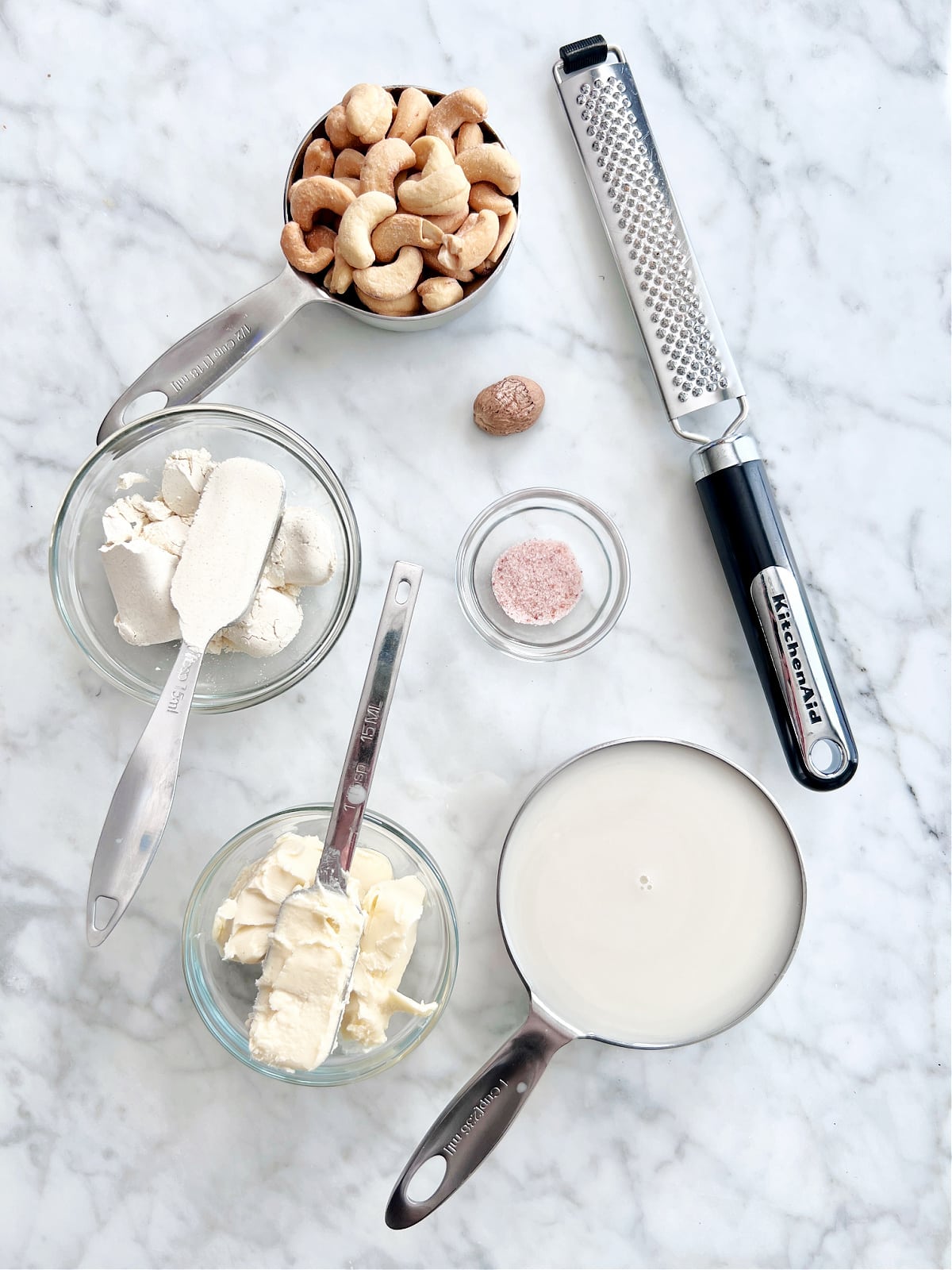 Overhead view of ingredients for cashew bechamel sauce: a measuring cup of cashews, a glass bowl of flour, a glass bowl of butter, a pinch bowl of sea salt, a measuring cup of dairy free milk, a nutmeg pod and grater.