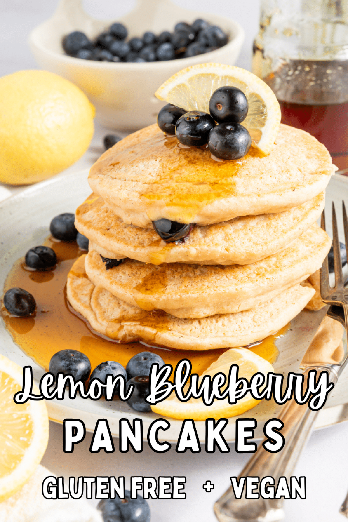 A stack of pancakes on a rustic white plate, garnished with fresh blueberries, lemon slices, and syrup. A fork and knife sit on the plate, and there is one triangle bite cut out of the pancake stack.