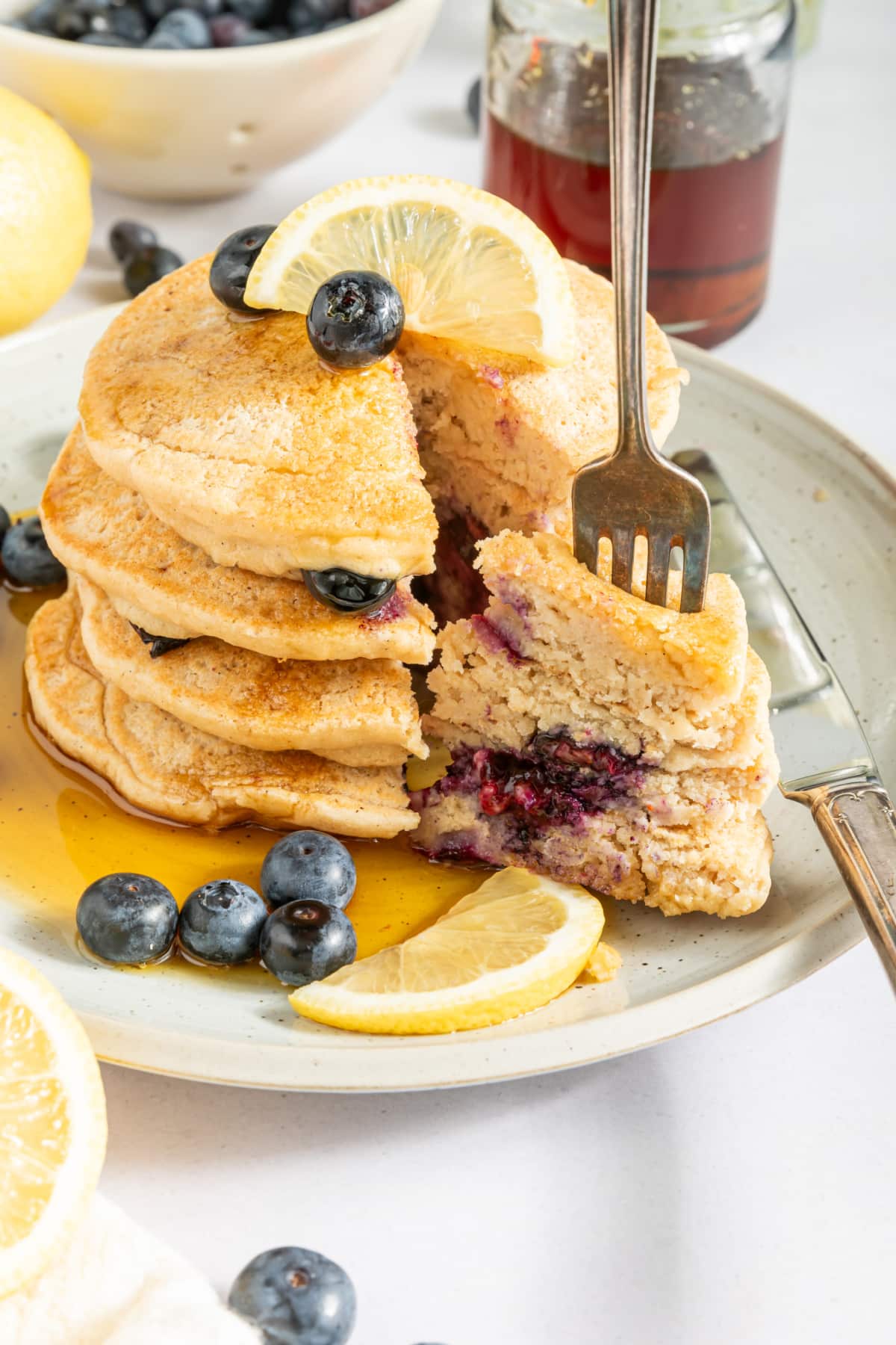 A stack of pancakes on a rustic white plate, garnished with fresh blueberries, lemon slices, and syrup. A knife sits on the plate, and there is one triangle bite cut out of the pancake stack, still on the plate with a fork in it.