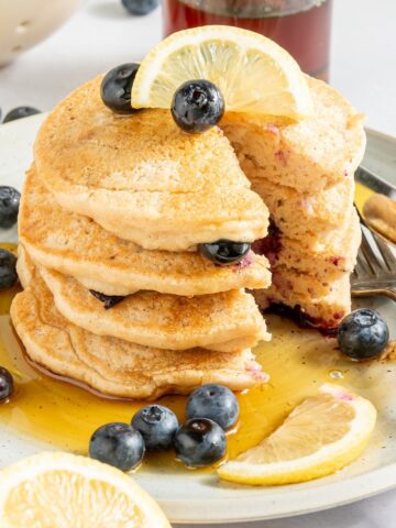 A stack of four lemon blueberry pancakes on a rustic white plate, garnished with fresh blueberries, lemon slices, and syrup. A fork and knife sit on the plate, and there is one triangle bite cut out of the pancake stack.