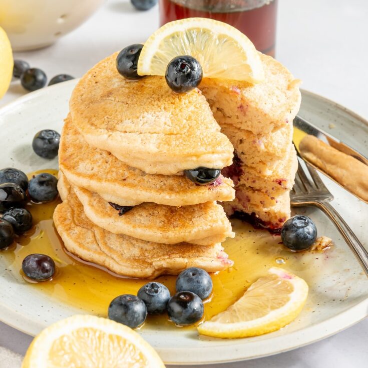 A stack of four lemon blueberry pancakes on a rustic white plate, garnished with fresh blueberries, lemon slices, and syrup. A fork and knife sit on the plate, and there is one triangle bite cut out of the pancake stack.