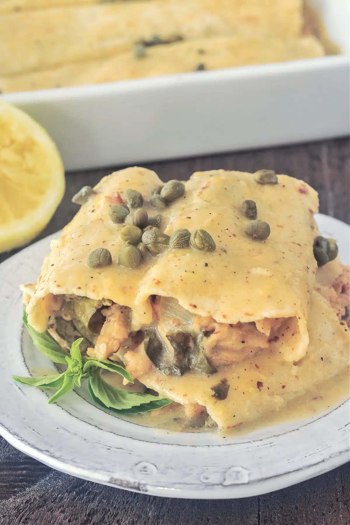 Close up of one serving of lemon piccata enchilada on a plate, next to a pan of enchiladas and a squeezed lemon half.