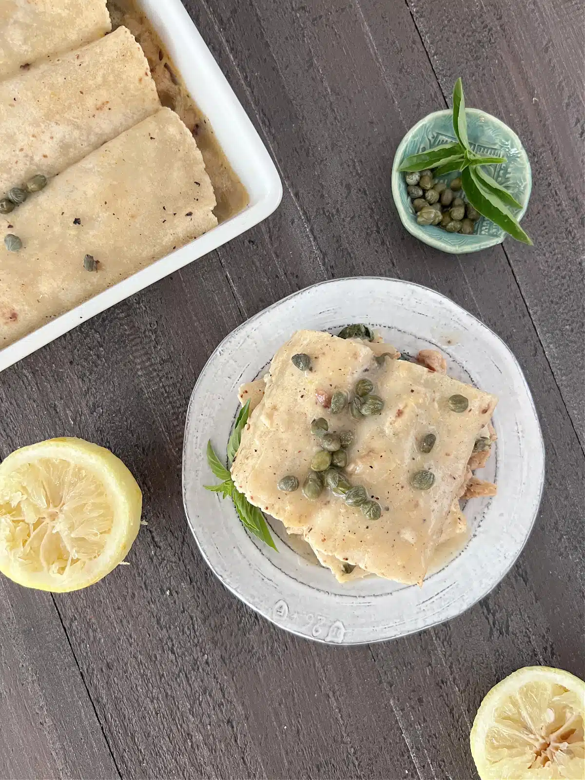 Overhead view of one serving of lemon piccata enchilada on a plate, next to a pan of enchiladas, a squeezed lemon half, and a small bowl of capers.