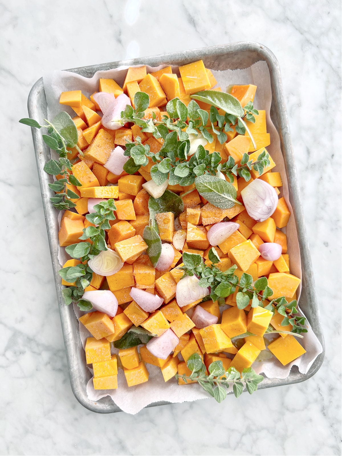 Overhead view of cubes of butternut squash, several garlic cloves and shallots, sprigs of oregano and sage leaves on a baking sheet ready to roast.