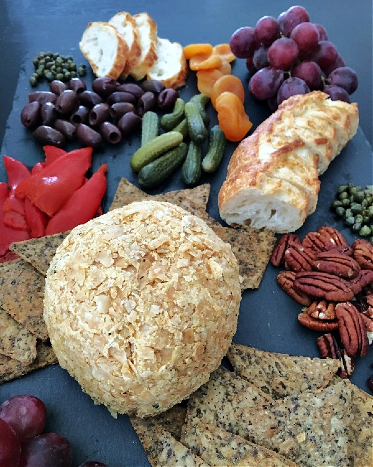 A pumpkin cheese ball on a black slate board with crackers, pecans, capers, sliced baguette, purple grapes, cornichon pickles, and olives.