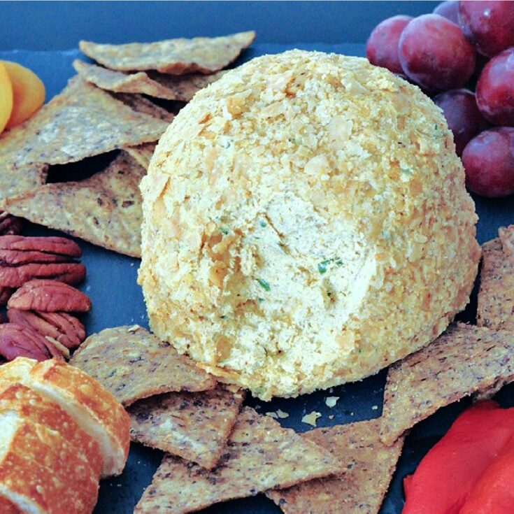 Close up of a golden brown colored spiced pumpkin basil cheese ball on a black slate board with crackers, pecans, capers, sliced baguette, purple grapes, cornichon pickles, and olives.