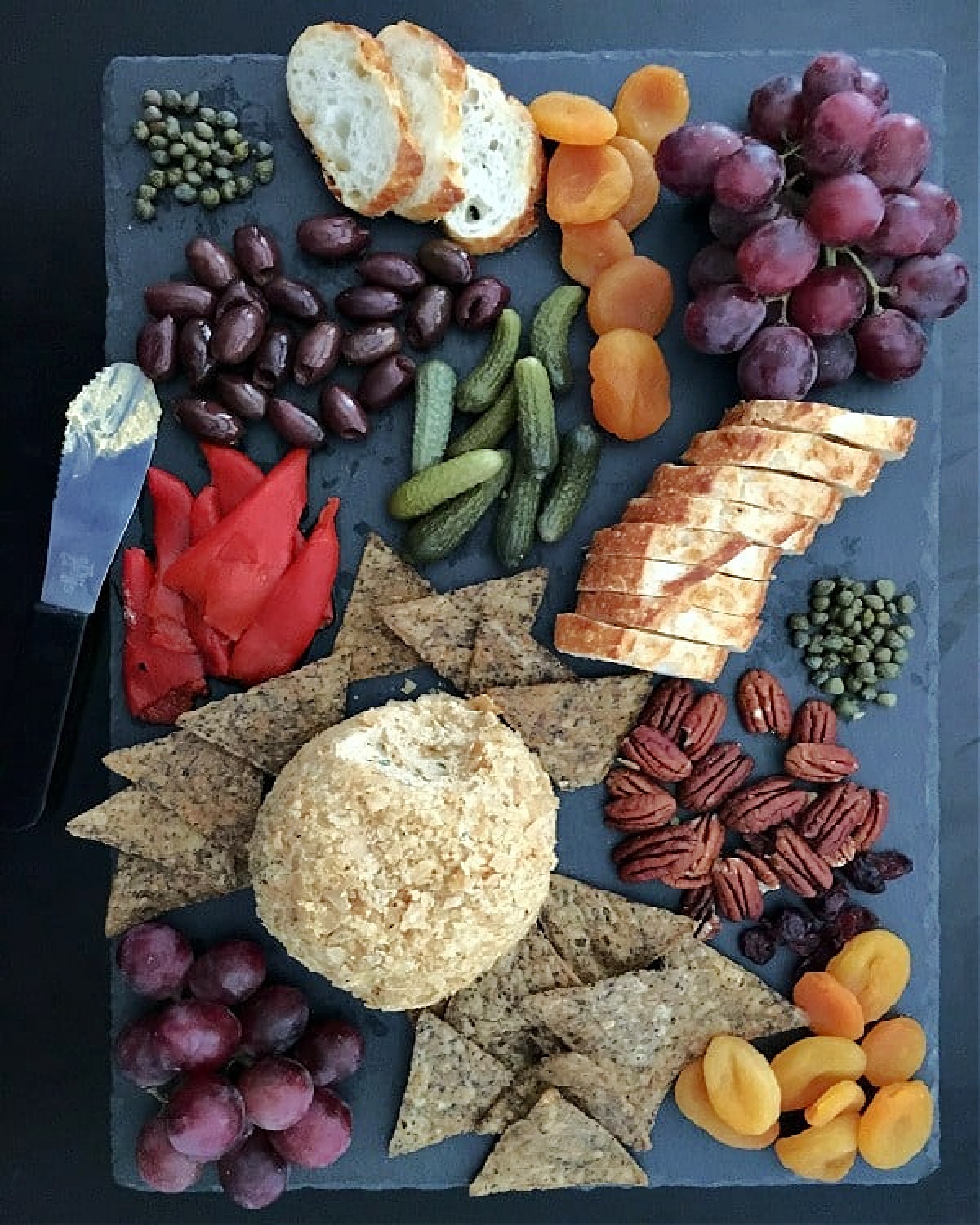 Overhead view of a golden brown colored spiced pumpkin basil cheese ball on a black slate board with crackers, pecans, capers, sliced baguette, purple grapes, cornichon pickles, and olives.