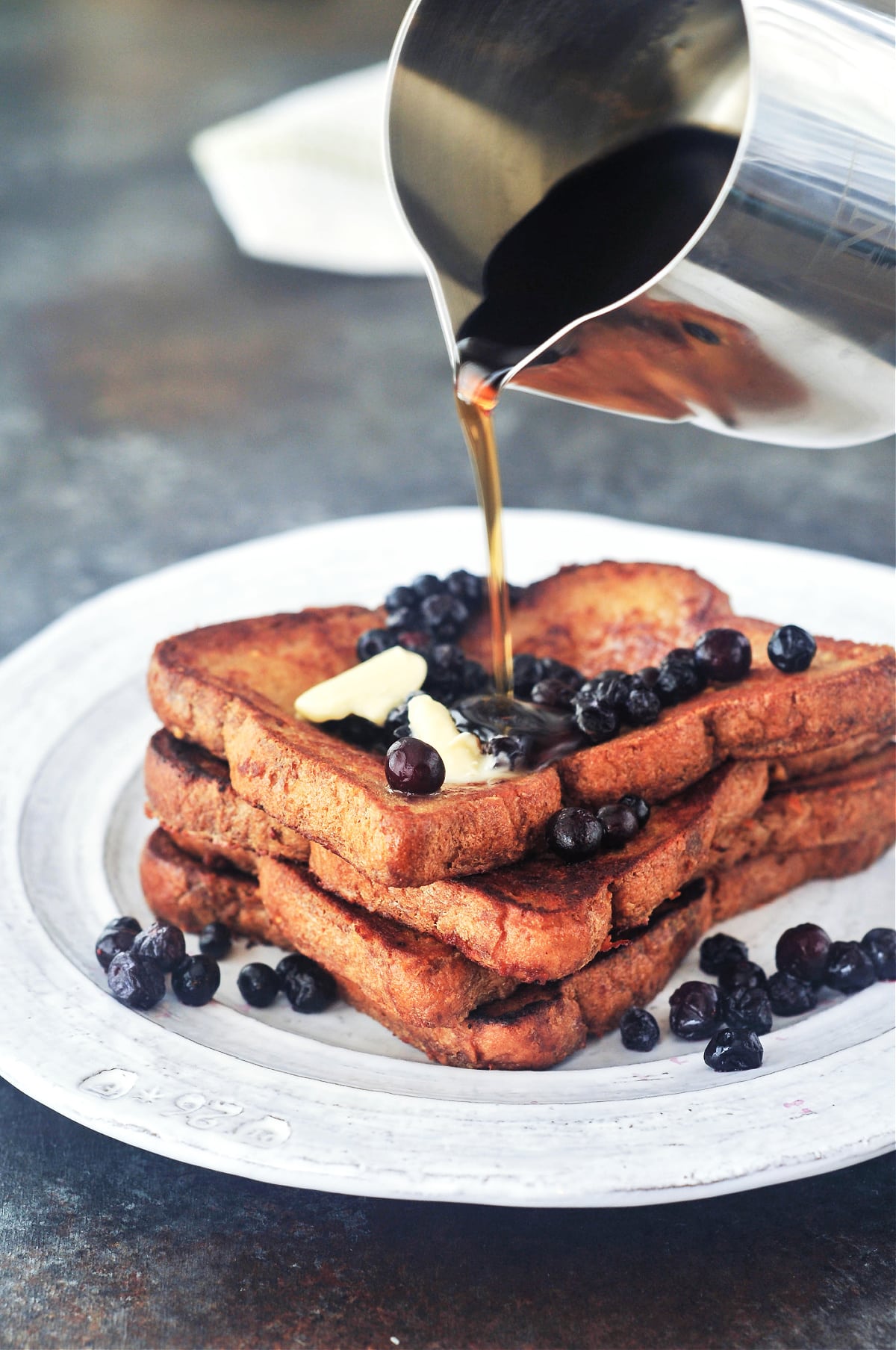 A stack of four slices of Baileys Irish cream French toast on a rustic white plate. French toast is garnished with blueberries and a silver pitcher pours maple syrup over the top.