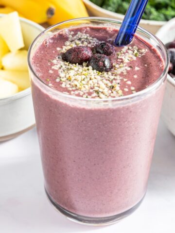 Purple blueberry smoothie in pint glass topped with frozen blueberries and hemp seeds, with a blue glass straw. Bowls of smoothie ingredients behind the glass: pineapple, a banana bunch, spinach, and blueberries.