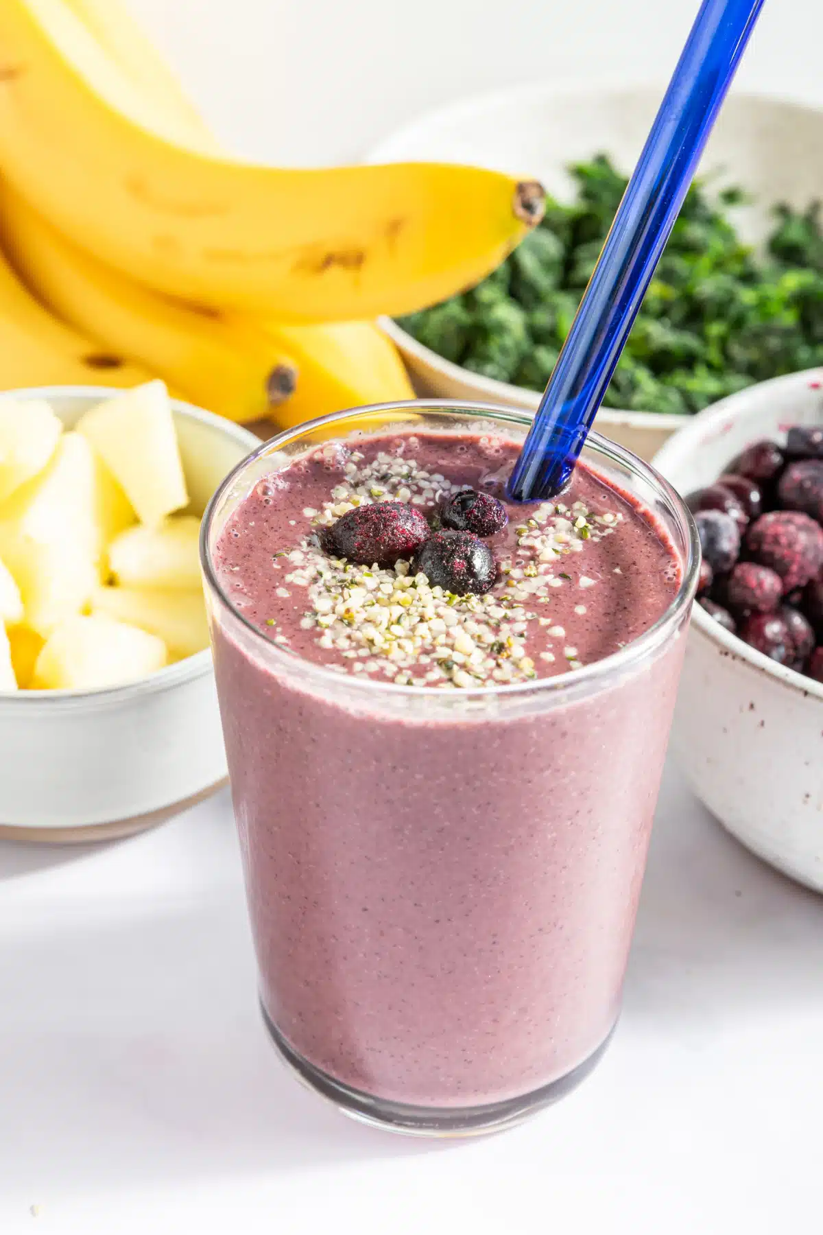 Purple blueberry smoothie in pint glass topped with frozen blueberries and hemp seeds, with a blue glass straw. Bowls of smoothie ingredients behind the glass: pineapple, a banana bunch, spinach, and blueberries.