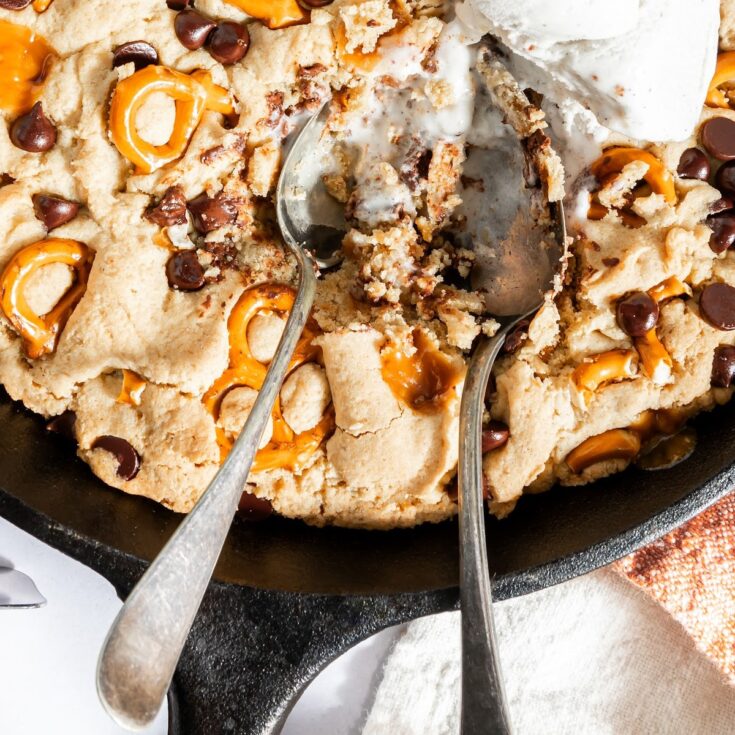 Overhead view of a baked skillet cookie with caramel, pretzels, and chocolate chips. Three scoops of vanilla ice cream melting on top, with two spoons digging into the cookie.
